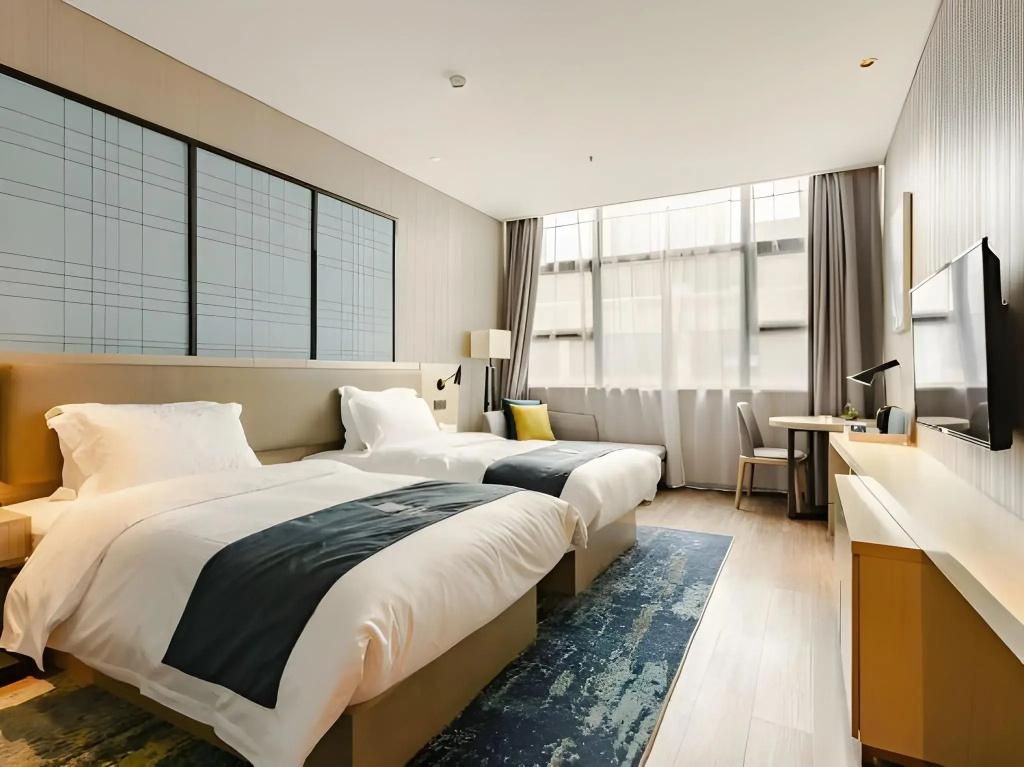 A hotel room with two beds, a desk, a chair, a TV, and large windows.