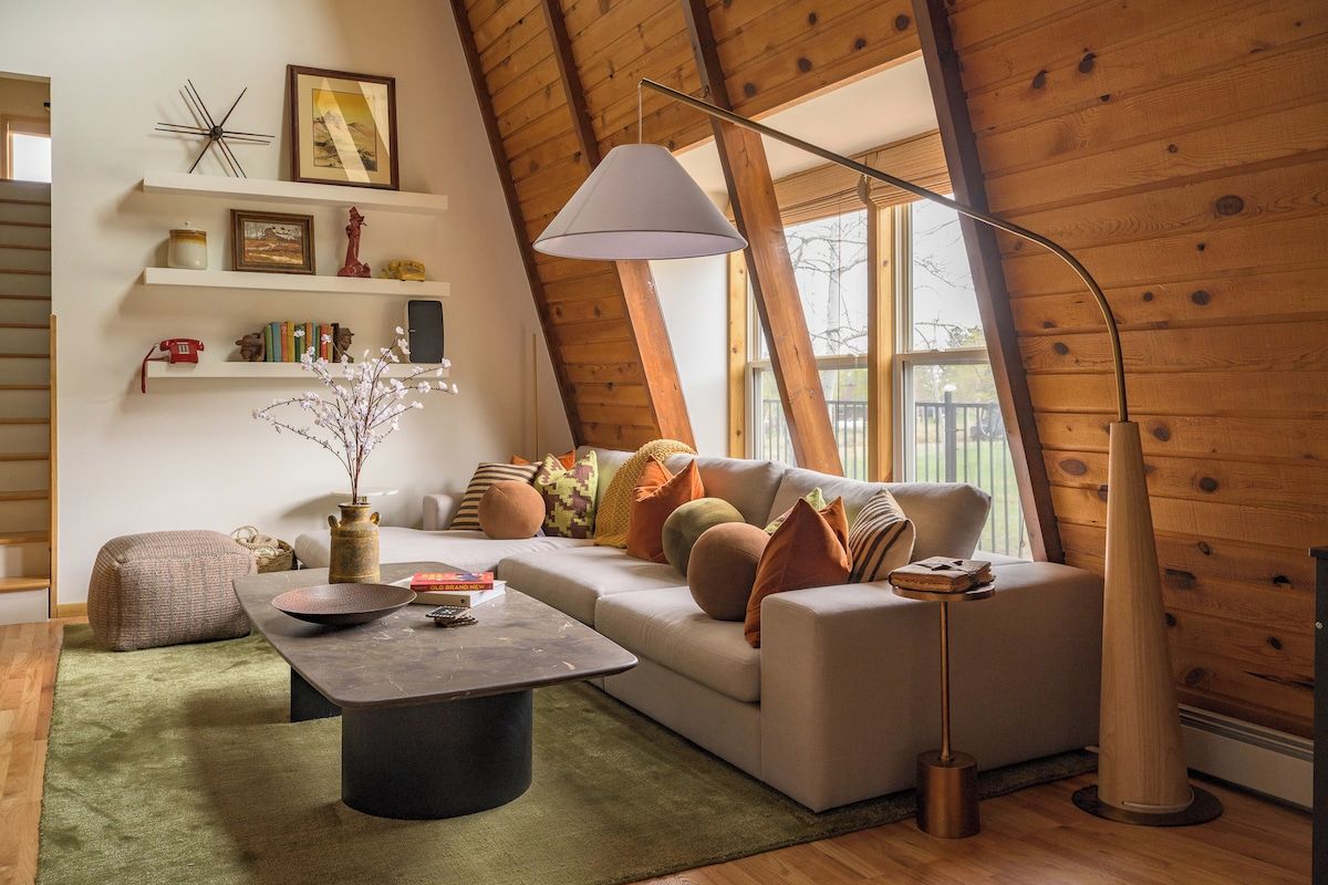 A cozy living room with a sofa, coffee table, and a lamp. There are shelves with decorative items and a window with a view.