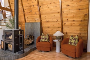 Una habitación acogedora con una estufa de leña, dos sillones con cojines estampados, una lámpara sobre una mesita auxiliar y una escalera con barandillas de madera.
