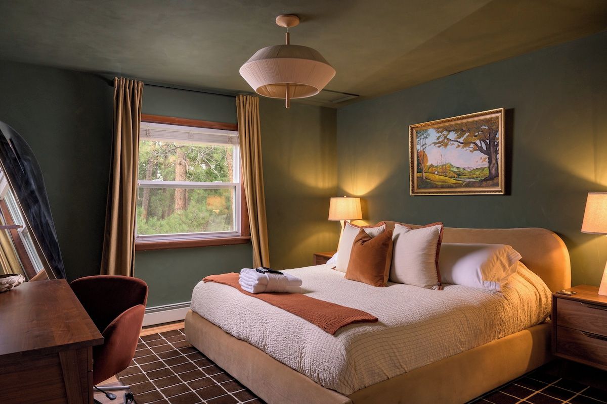 A hotel room with a bed, bedside lamps, a desk, a chair, a window with curtains, and a painting on the wall.
