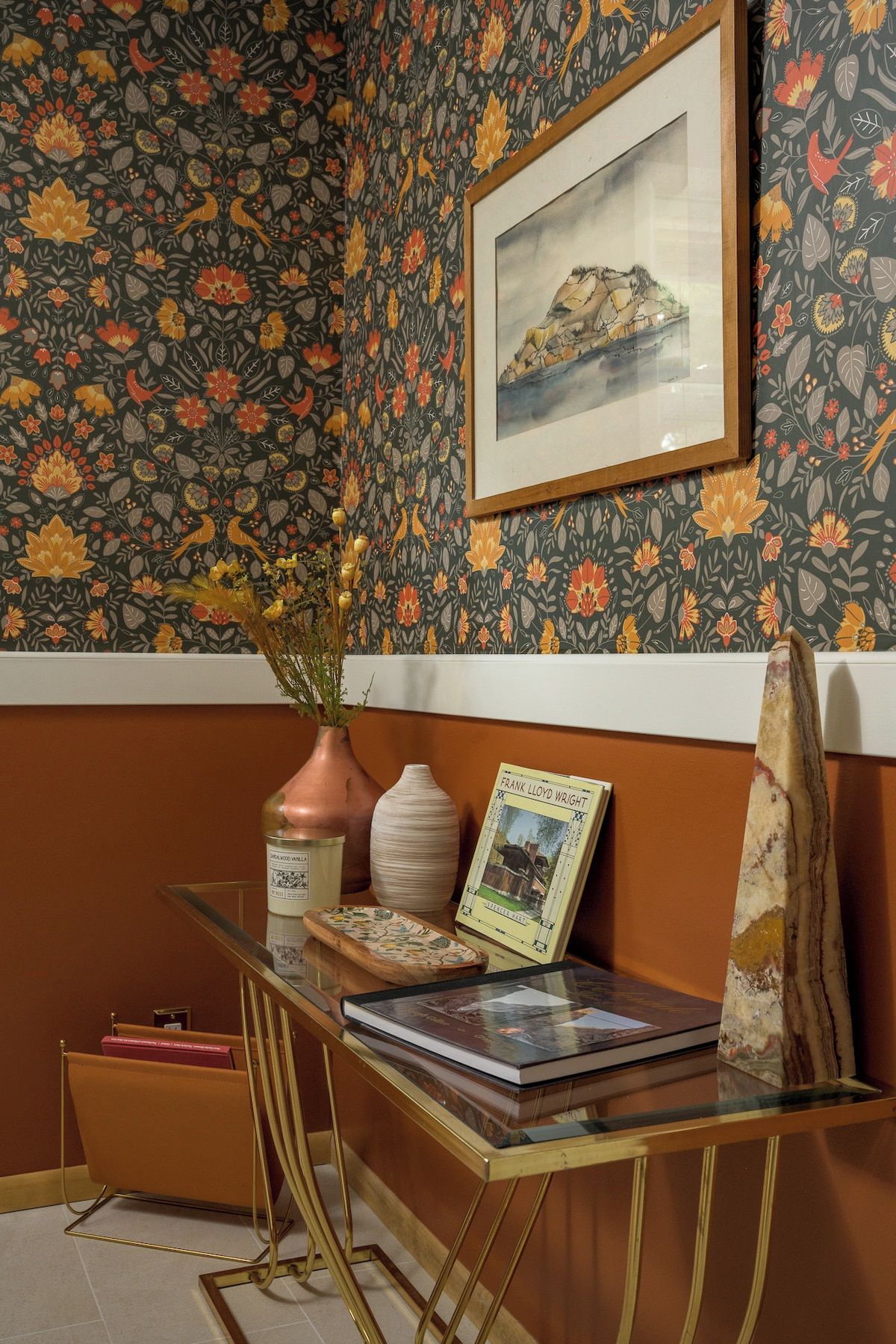 A room with a patterned wallpaper, a framed picture on the wall, a glass table with a vase, a book, and a decorative object.