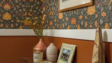 A room with a patterned wallpaper, a framed picture on the wall, a glass table with a vase, a book, and a decorative object.