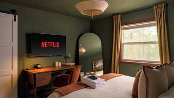 Ett hotellrum med en säng, ett skrivbord med en dator, en tv som visar ”NETFLIX”, ett fönster med gardiner och en lampa.