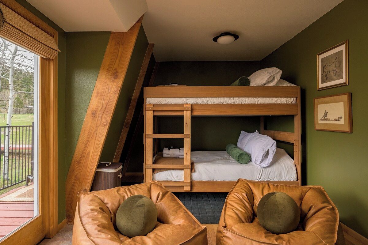 A dorm-style room with bunk beds, a window with blinds, a small table, and two leather chairs.