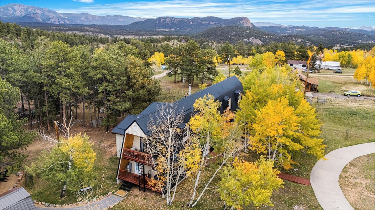 A two-story cabin with a wrap-around porch, surrounded by a forested area with autumn foliage, and a mountain range in the background.