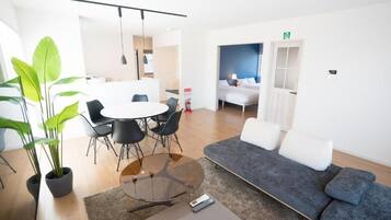 A modern hotel room with a sofa, a round table with chairs, a potted plant, and a bed with a view.