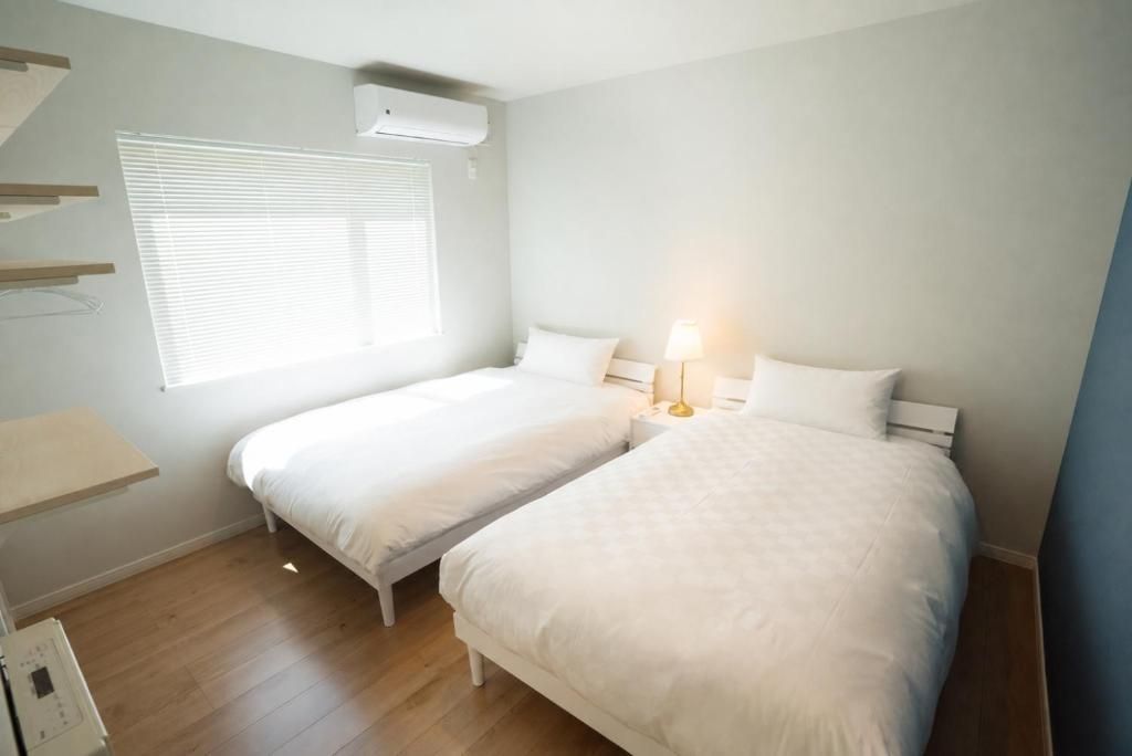 A bedroom with two beds, a bedside table with a lamp, a window with blinds, and a wall-mounted air conditioner.