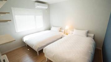 A bedroom with two beds, a bedside table with a lamp, a window with blinds, and a wall-mounted air conditioner.