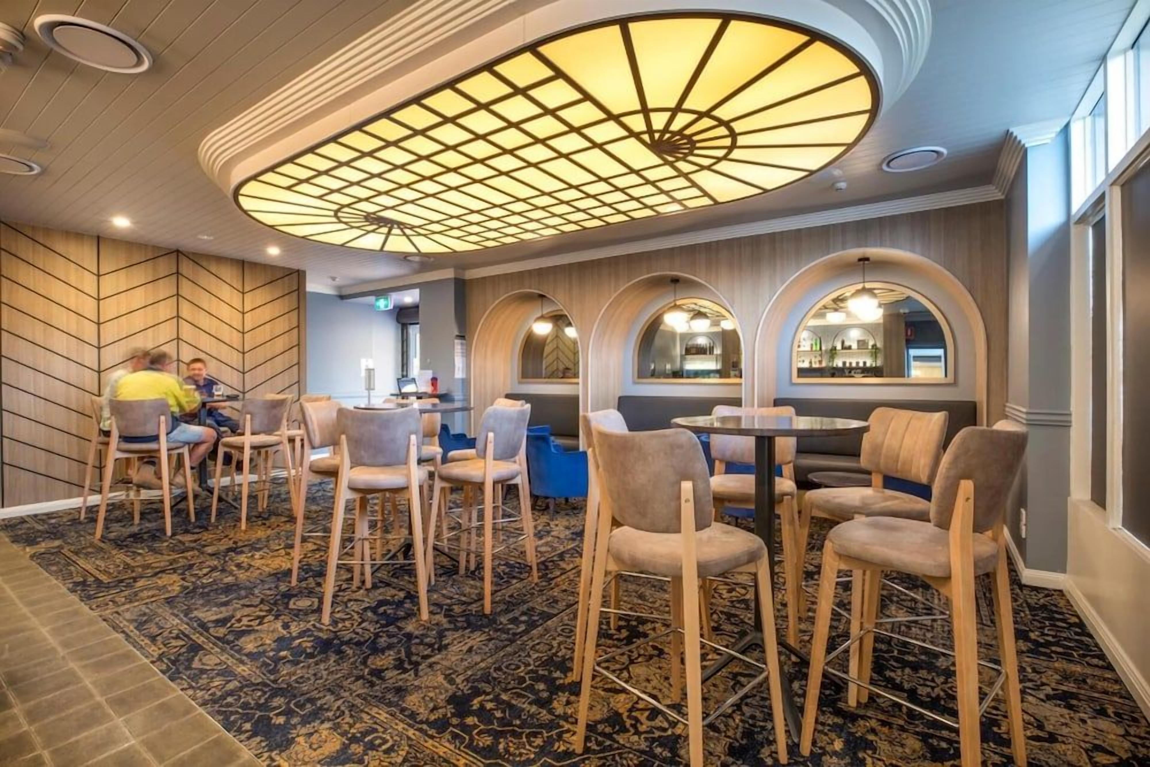 A hotel lobby with a patterned carpet, wooden chairs, a round table, and a ceiling with a geometric light fixture.