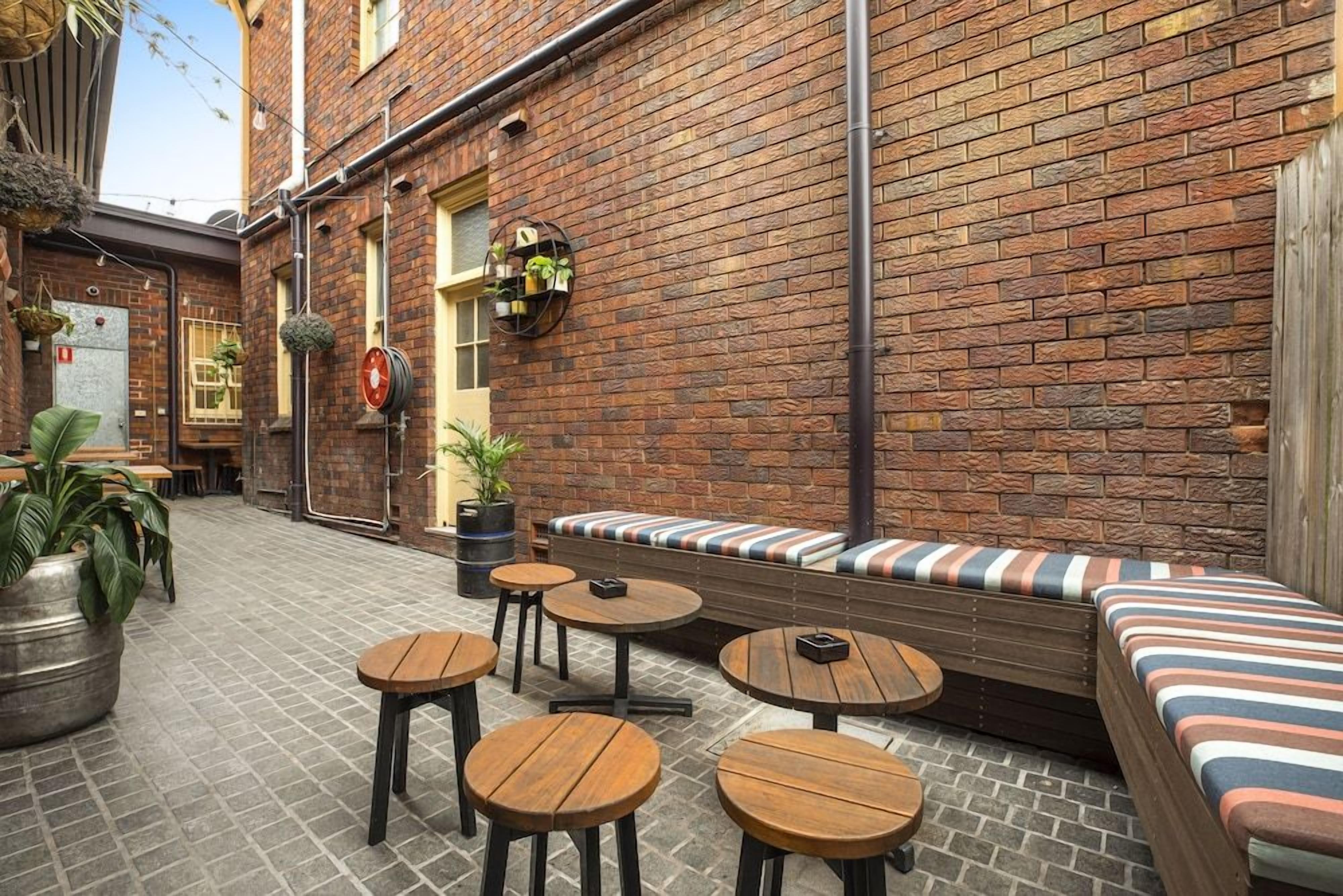 An outdoor seating area with round wooden tables and benches, a brick wall, and hanging planters.