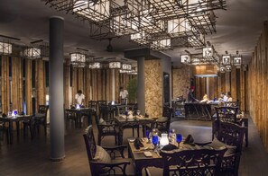An indoor dining area with bamboo walls, tables with blue glassware, and a chef cooking at a central island. - Emerald Maldives Resort and Spa Cache Transfer Included (Fasmendhoo)