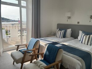 Habitación de hotel con dos camas, zona de estar con sillas y vista a la playa a través de amplias ventanas.