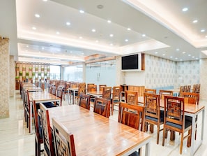 A spacious dining area with multiple wooden tables and chairs, a flat-screen TV mounted on the wall, and a patterned wall. - Suite Palette Patel Nagar (Bhopal)