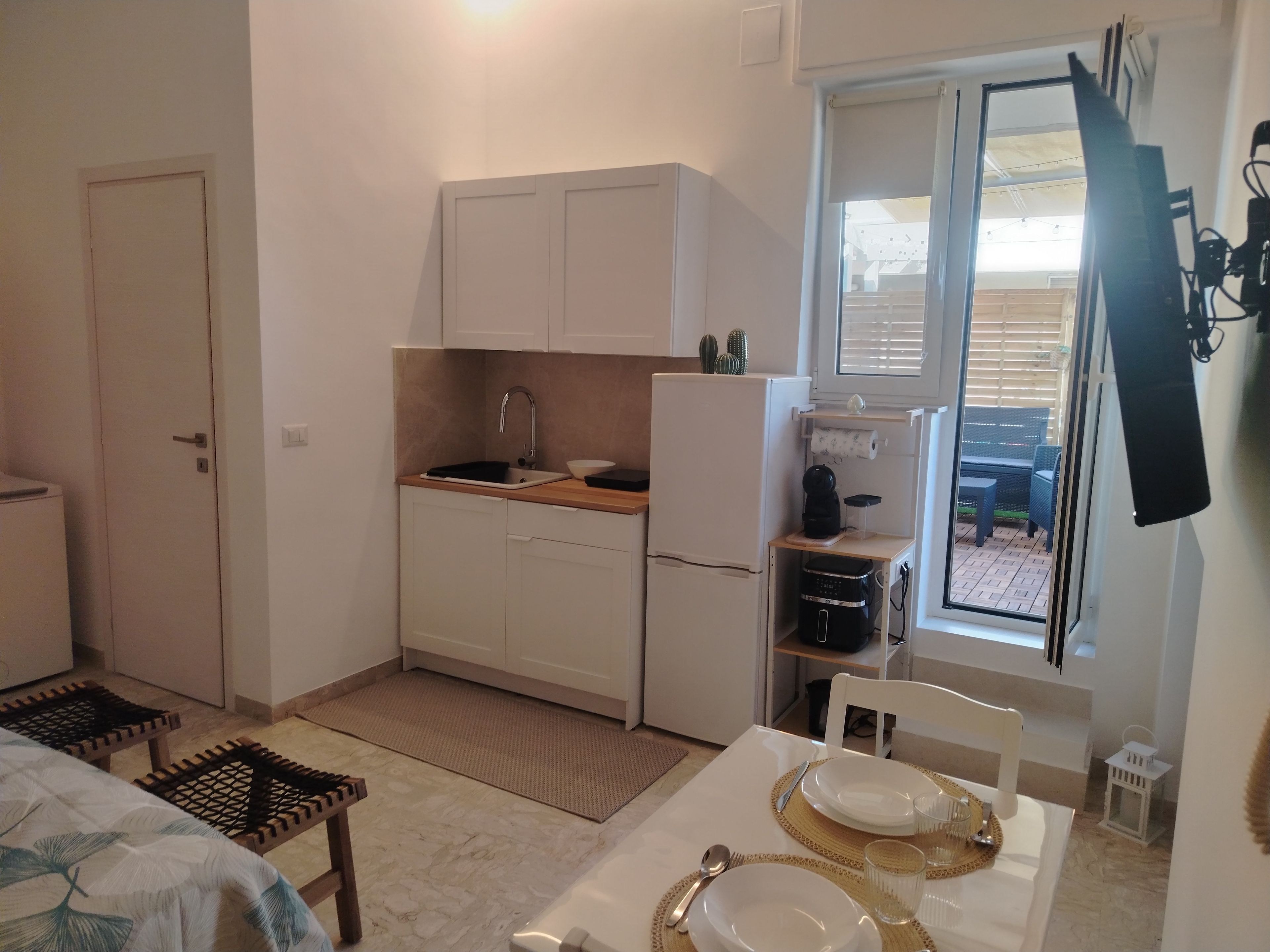 Una cucina compatta con mobili bianchi, un lavello e un frigorifero. È presente un tavolo da pranzo apparecchiato con piatti, posate e bicchieri.