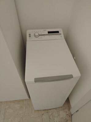 Lavadora blanca de carga frontal, compacta, ubicada en un rincón de una habitación con piso de baldosas.