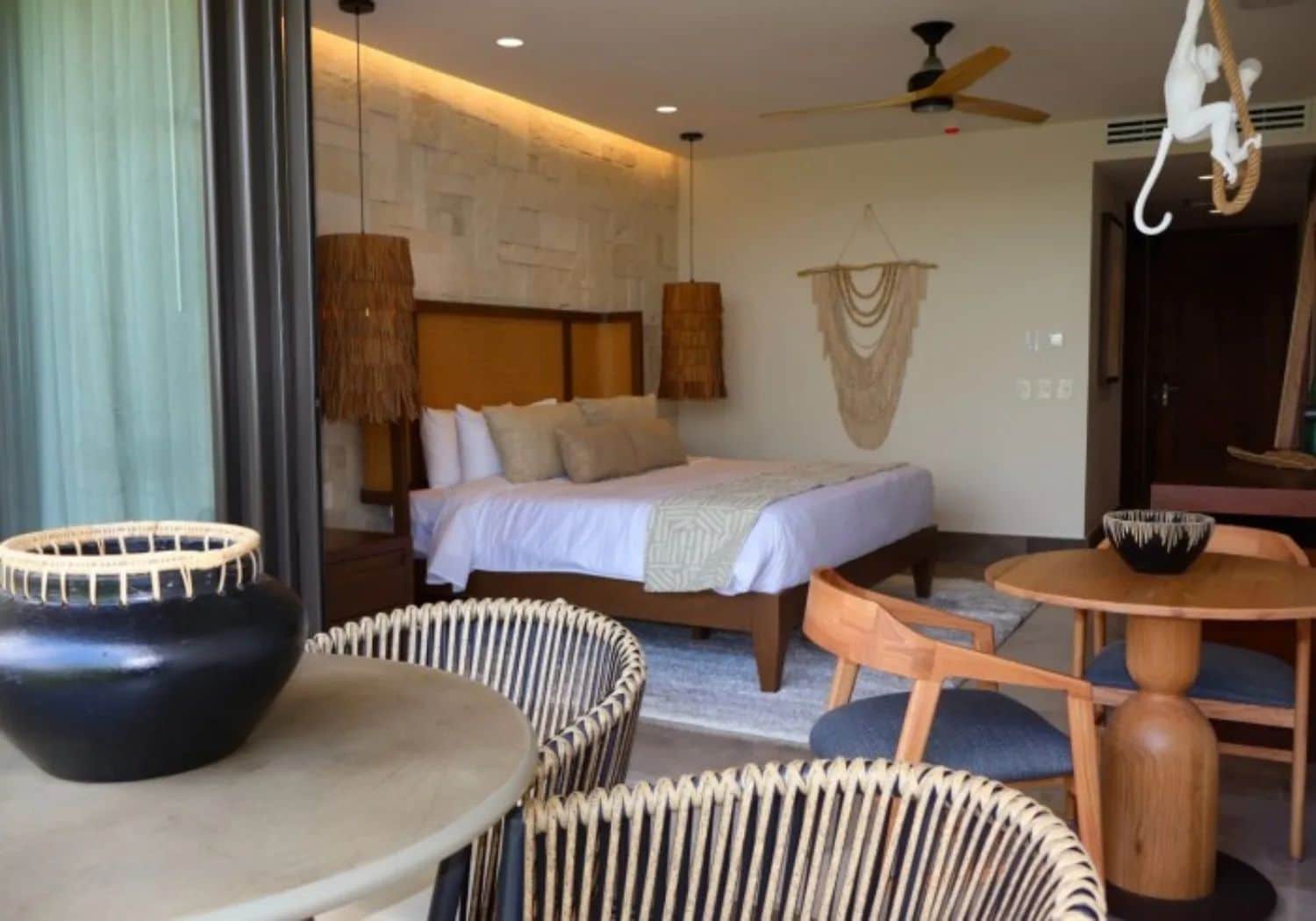 Una habitación de hotel con una cama, una mesa redonda de madera, sillas de mimbre, un tazón negro grande, un ventilador de techo y una lámpara de pared.