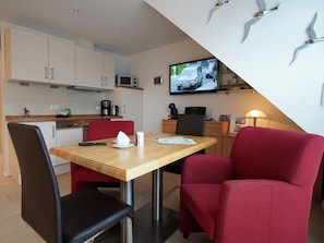 A compact hotel room with a kitchenette, a dining area with a table and chairs, a television mounted on the wall, and a red upholstered chair. - Holiday Apartment With 1 Bedroom (Norden)