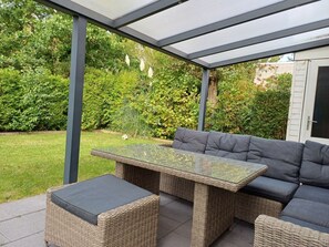 An outdoor patio area with a glass-top table, wicker furniture, and a view of a garden. - Strand4you Zeeland Village (Scharendijke)