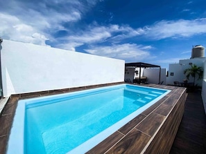 Ein rechteckiger Swimmingpool mit einer ihn umgebenden Holzterrasse, vor einer weißen Wand und unter blauem Himmel.