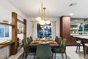 A modern dining room with a wooden table set for six, surrounded by green upholstered chairs. - Pool + Hot Tub + Game Room | Mtn Views + Fire Pit | The Royal Palms (Palm Springs)