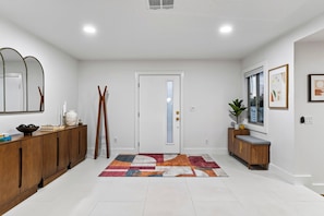 A modern hallway with a wooden cabinet, a mirror, a door with frosted glass, a patterned rug, and a small plant. - Pool + Hot Tub + Game Room | Mtn Views + Fire Pit | The Royal Palms (Palm Springs)