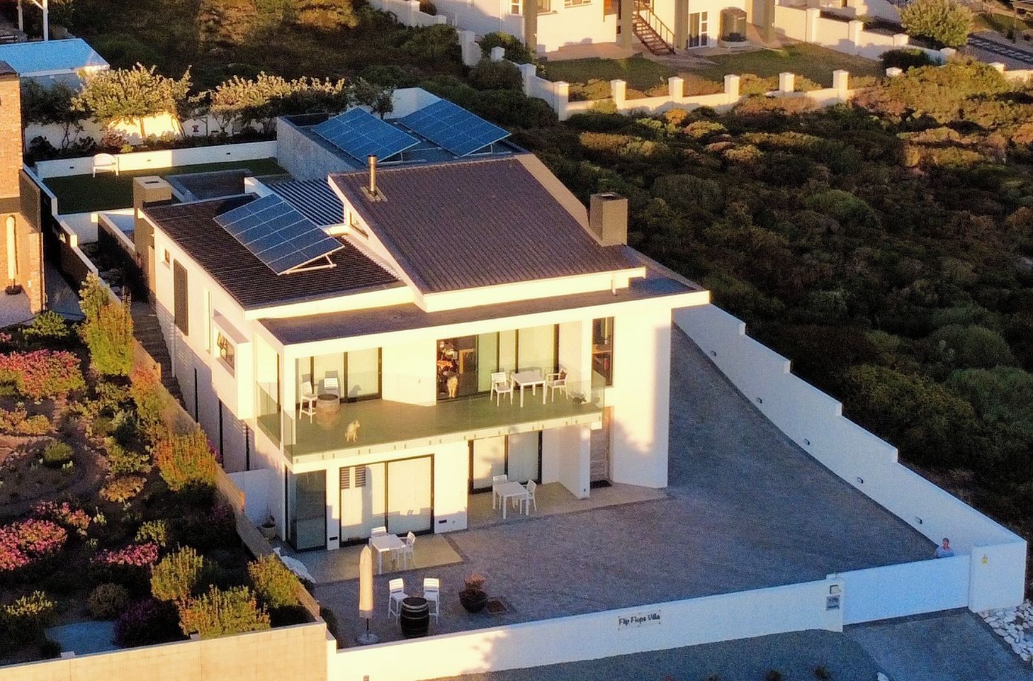 Une maison moderne à deux étages, dotée d’un toit plat, de panneaux solaires et d’un balcon avec un espace salon extérieur.
