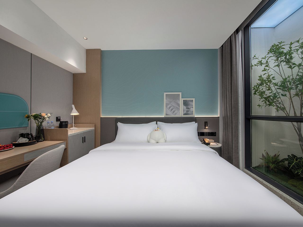 A modern hotel room with a large bed, a desk with a chair, a nightstand, and a window with a view of greenery.