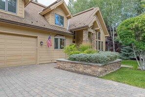 A two-story house with a garage, a stone planter with shrubs, and a flag. - Spacious Warm Springs Home | Hot Tub + Bunk Room (Ketchum)