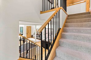 A staircase with wooden handrails and black balusters, leading to a landing with a view of a living room. - Spacious Warm Springs Home | Hot Tub + Bunk Room (Ketchum)
