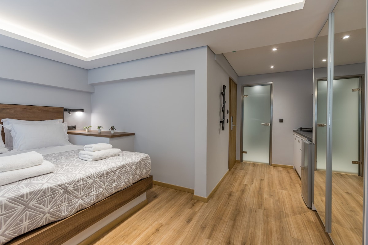 Ein modernes Hotelzimmer mit einem hölzernen Bett, einem Nachttisch, einem integrierten Schreibtisch, einem Spiegel und einem Flur, der zum Badezimmer führt.