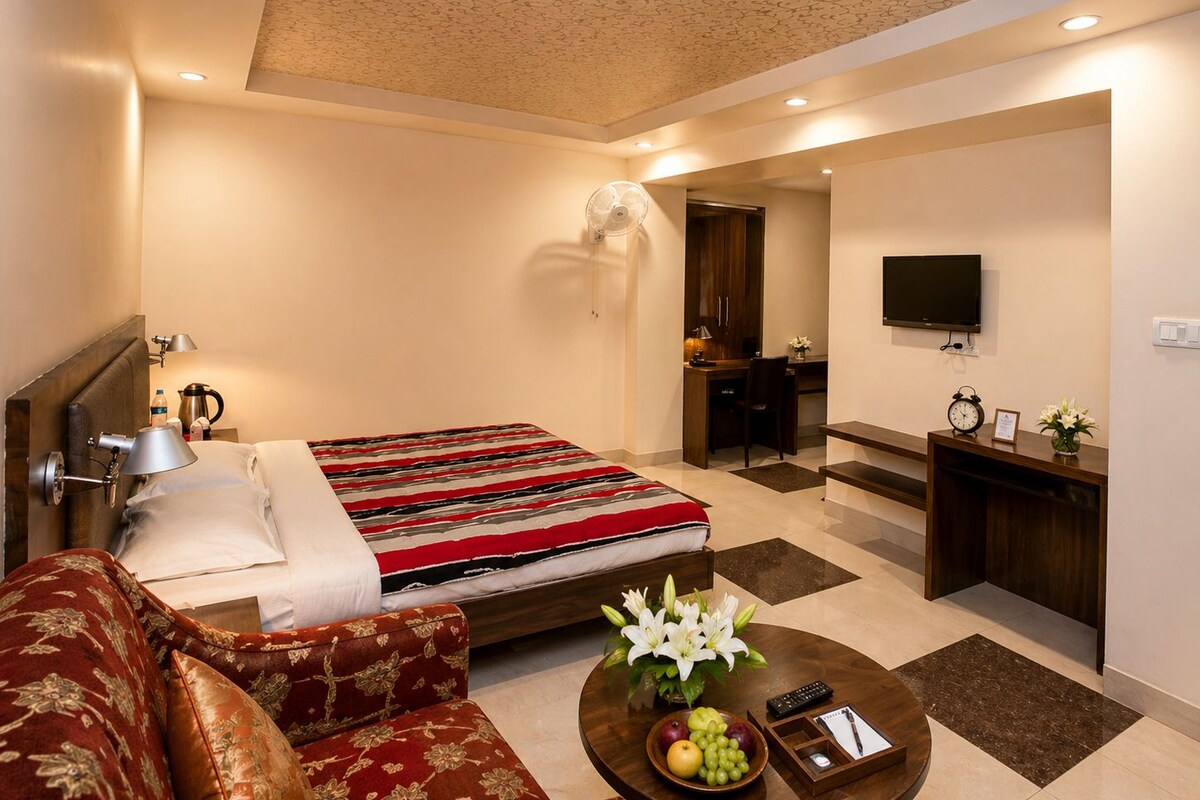 A hotel room with a bed, a sofa, a desk, a TV, and a coffee table with fruits and flowers.