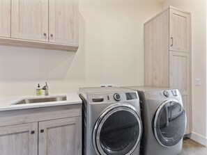 A laundry area with two front-loading washing machines, a sink with soap dispenser, and wooden cabinets. - New ! Big Sky Basin Home with Hot Tub, Game Room & Mountain Views (Big Sky)
