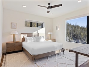 A bedroom with a bed, two bedside tables, a ceiling fan, and a large window with a view of trees. - New ! Big Sky Basin Home with Hot Tub, Game Room & Mountain Views (Big Sky)