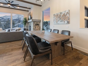 A modern dining area with a wooden table and leather chairs, a stone fireplace, and a large window with a mountain view. - New ! Big Sky Basin Home with Hot Tub, Game Room & Mountain Views (Big Sky)