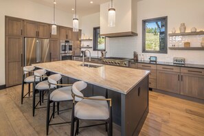A modern kitchen with a large island, stainless steel appliances, and a window overlooking a garden. - New! Entire Home Big Sky Summit - Private Hot Tub with Ski-In Ski-Out Access (Big Sky)