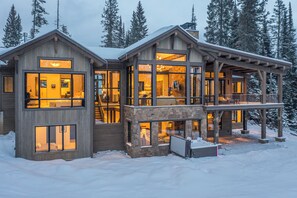 Een ruime, meerlaagse houten hut met grote ramen en een balkon, omgeven door sneeuw en bomen.