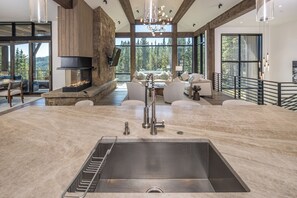 A spacious kitchen with a large island, a built-in fireplace, and a view of the forest through large windows. - New! Entire Home Big Sky Summit - Private Hot Tub with Ski-In Ski-Out Access (Big Sky)