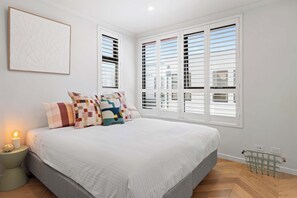 A bedroom with a large bed, two windows with shutters, a bedside table with a lamp, and a wall-mounted artwork. - Waterline Rise - Bay Views Williamstown (Williamstown)