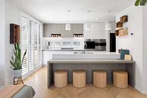 A modern kitchen with a central island, bar stools, and a large window with blinds. - Waterline Rise - Bay Views Williamstown (Williamstown)