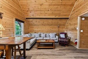 Ein gemütliches Blockhauss-Interieur mit Holzbalkendecke, einer Couch, einem gemusterten Sessel und einem Esstisch.
