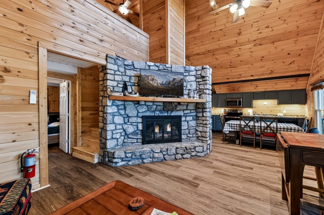 A log cabin interior with a stone fireplace, wooden walls, a dining area, and a kitchen.