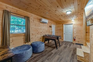 Une pièce chaleureuse avec des murs en bois, une table de baby-foot, deux poufs, une fenêtre avec des rideaux et une porte.