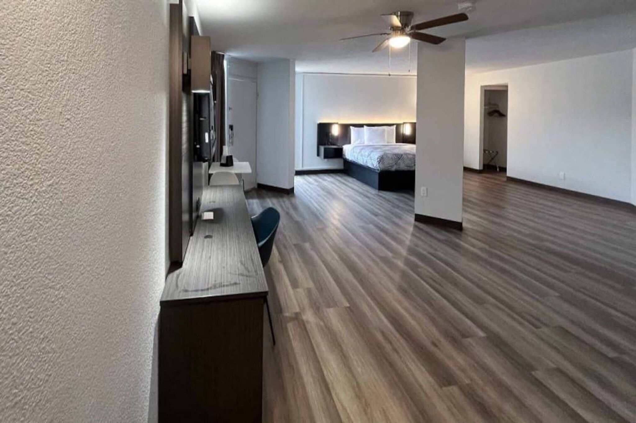 Een ruime kamer met een houten vloer, een bed, een bureau en een plafondventilator.