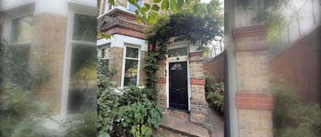 A brick building with a black door, a window, and a small garden area with green foliage.