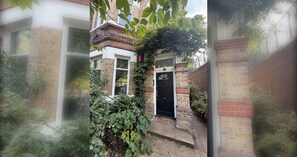A brick building with a black door, a window, and a small garden area with green foliage. - Characterful 1BDR Flat with Desk in Brixton Hill (London)