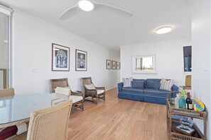 A living room with a glass-top dining table, wicker chairs, a blue sofa, and a ceiling fan. - Beachfront Apt w/ Pool & Spectacular Lake Views (Forster)