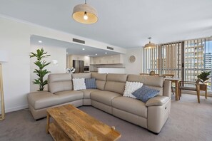 A modern living room with a sectional sofa, a wooden coffee table, and a dining area with a view of the city. - 'Breeze' at The Crest - Spacious & Secure Parking (Forster)