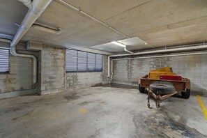 A concrete parking garage with a wheelie bin, a closed shutter door, and visible pipes on the ceiling. - 'Breeze' at The Crest - Spacious & Secure Parking (Forster)