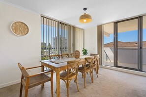 Un coin repas avec une table en bois, des chaises en osier, un luminaire en fibres tressées et une vue sur la ville à travers de grandes fenêtres.
