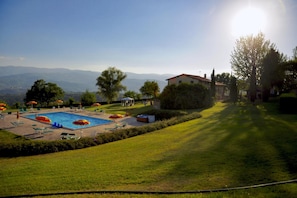 A swimming pool with lounge chairs and umbrellas, surrounded by a well-maintained lawn and a building with a red roof. - Tuscan Apartment with Pool & Views (Poppi)
